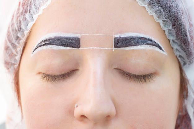 A close-up view of a woman's face with her eyes closed, displaying freshly microbladed eyebrows with measurement markings to ensure symmetry. She is wearing a protective hair net and a small nose piercing is visible.