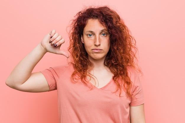 Red-haired woman giving a thumbs down gesture against a pink background