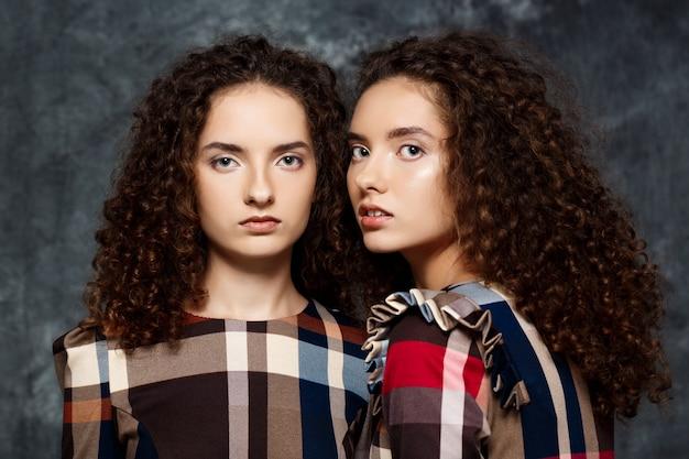 Two women with curly hair wearing plaid dresses standing against a textured blue background