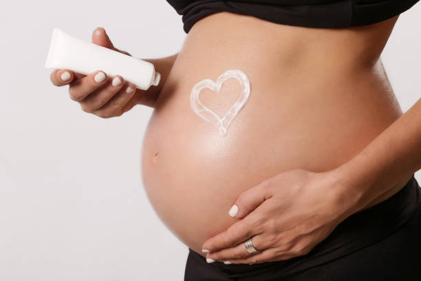 Pregnant woman applying lotion on belly with heart shape.